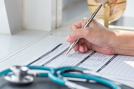 Doctor writing on medical health care record, patients discharge, or prescription form paperwork in hospital clinic office with physician’s stethoscope on desk